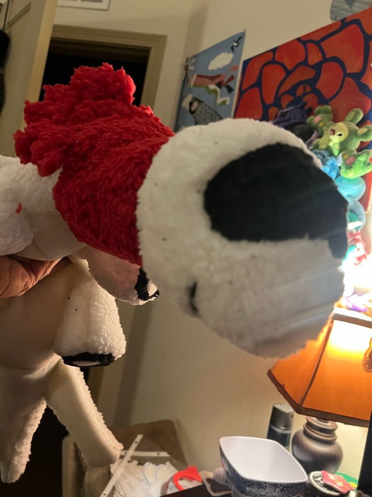 A plushie of a dragon designed to look like an airplane made with fluffy white and black fabric. It has wheels on the bottoms of its feet, and a bright red scarf around its neck