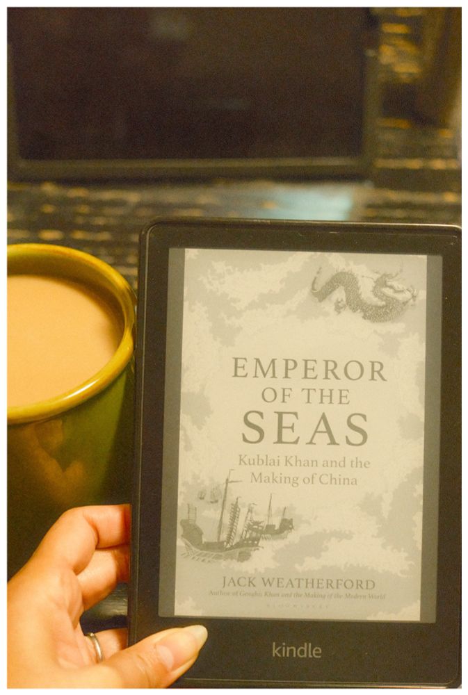 a Kindle showing the book cover “Emperor of the Seas: Kublai Khan and the Making of China” beside a mug of coffee on a table.