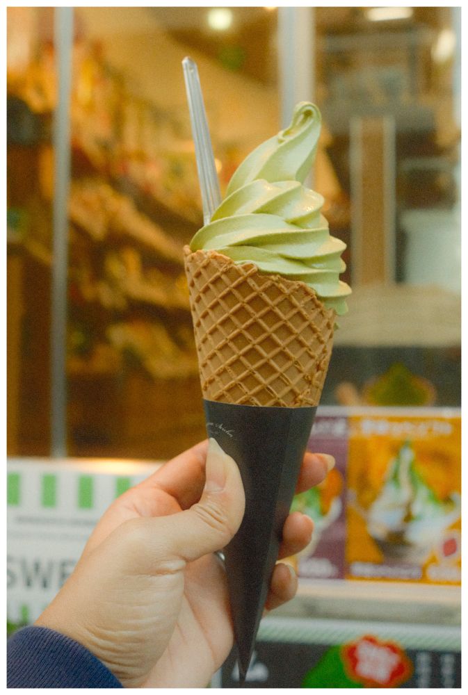 Close-up of a hand holding a tall waffle cone filled with swirled green matcha soft serve, a small clear spoon tucked into the side, with the warm, slightly blurred interior of a shop in the background.