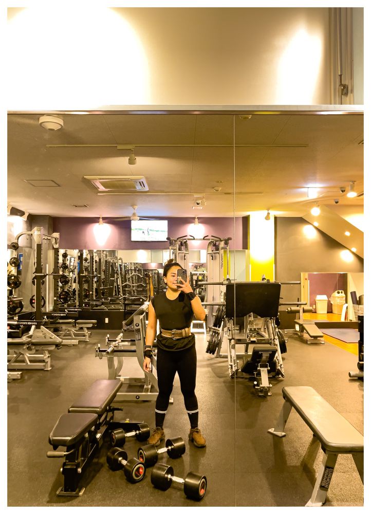 Mirror selfie in a mostly empty gym: a person in black workout clothes with a lifting belt stands between benches and weight machines, with several dumbbells on the floor in front of them.