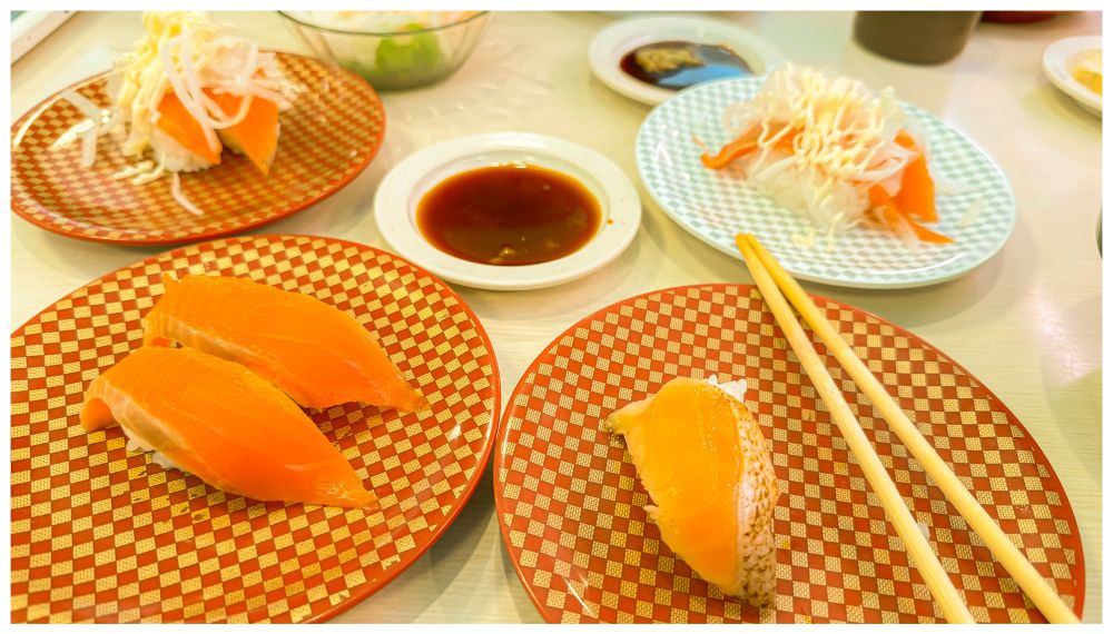 Plates of salmon nigiri and salmon sashimi topped with shredded onion and mayo on a restaurant table, with soy sauce dishes and wooden chopsticks nearby.