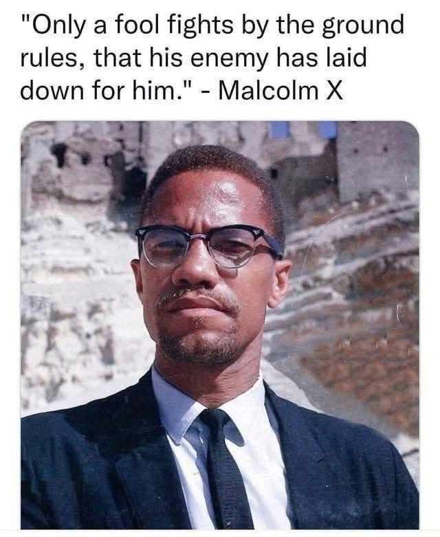 A color photo portrait of Malcolm X standing in front of what looks like the ruins of a castle. He is wearing an austere black suit and tie and glasses with browline black frames. The Sun is shining. The text says, "Only a fool fights by the ground rules that his enemy has laid down for him." -Malcolm X.