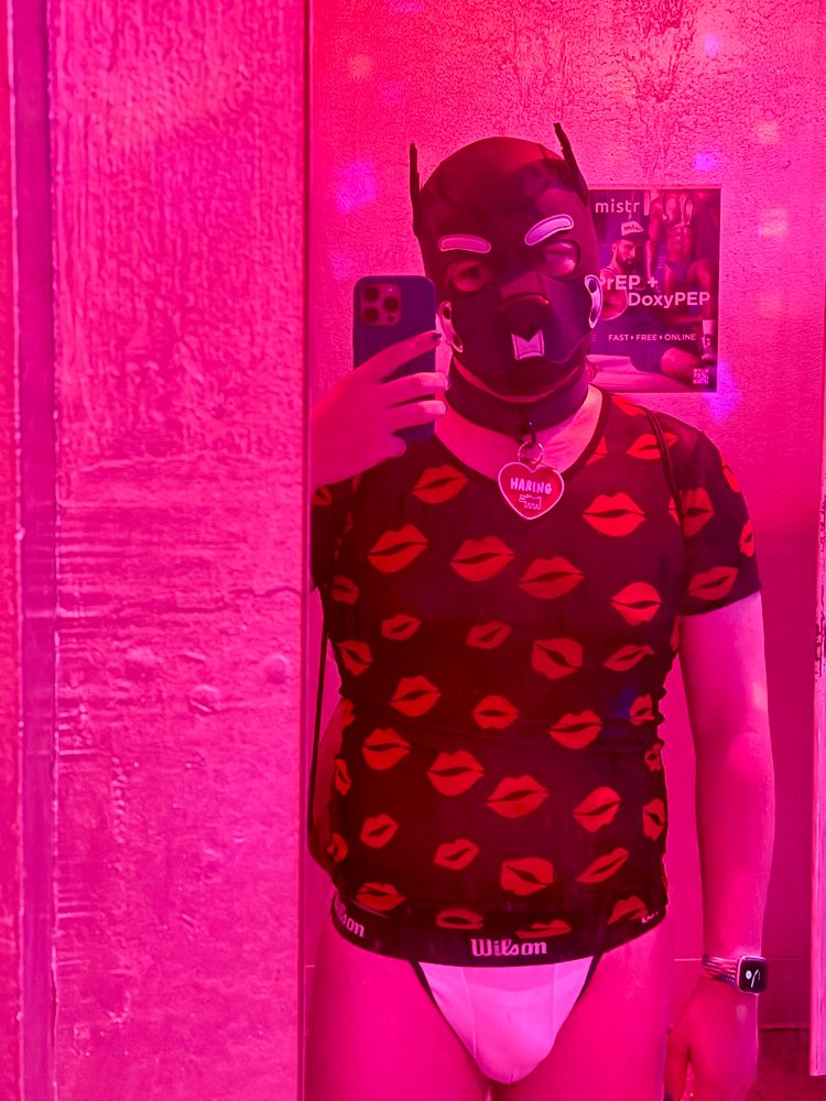 Haring standing in front of the mirror of a gay bar, with pink lighting. They are wearing a mesh top with red kiss marks all over it, and a white Wilson jockstrap. Their tag is the red Haring tag used for Valentine’s Day 2025.
