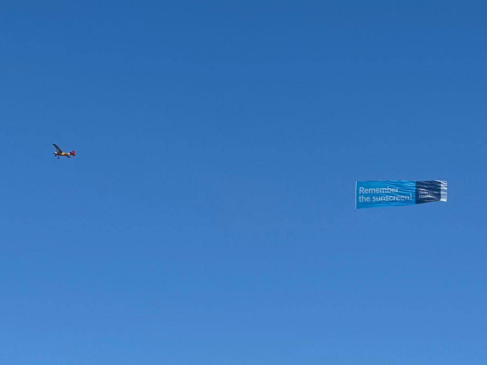 A plane in a clear cloudless blue sky towing a banner that reads: “Remember the sunscreen” on a light blue field with “Yale New Haven Health” on a darker blue field on the end. 