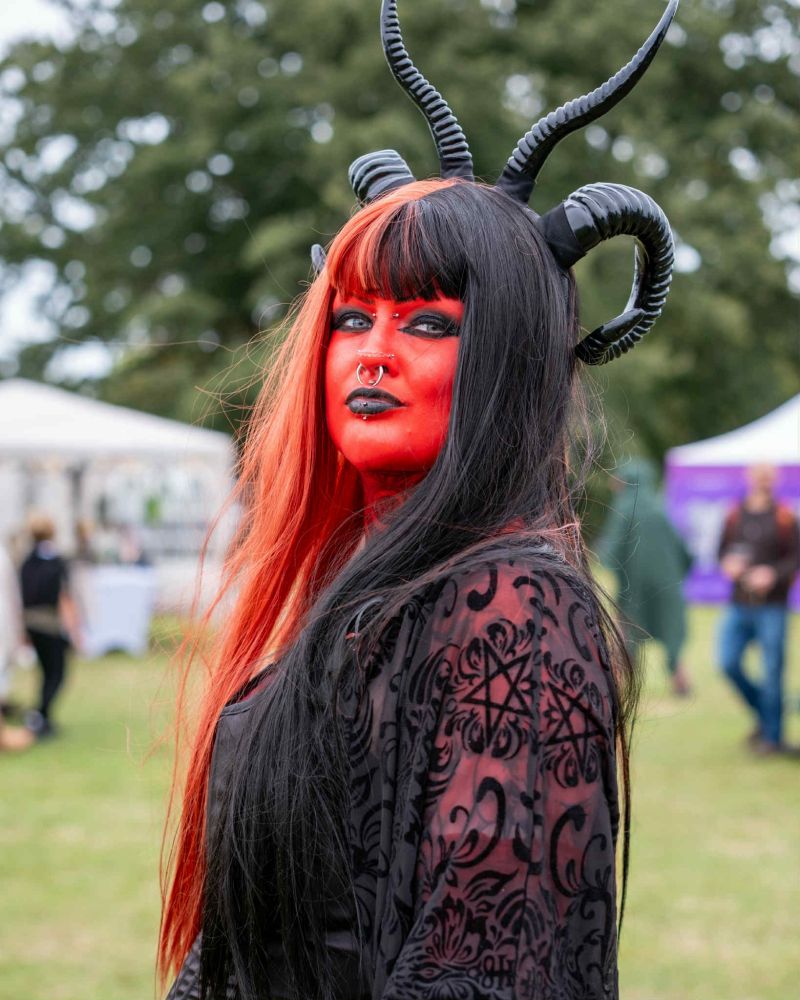 This is a photo of Mx River. They are stood facing the side, with their head facing the camera. It is cropped at their chest. Mx River is dressed as a Tiefling, with their skin painted red, black horns, a corset, harem style pants, creeper shoes and a belt with potions on.