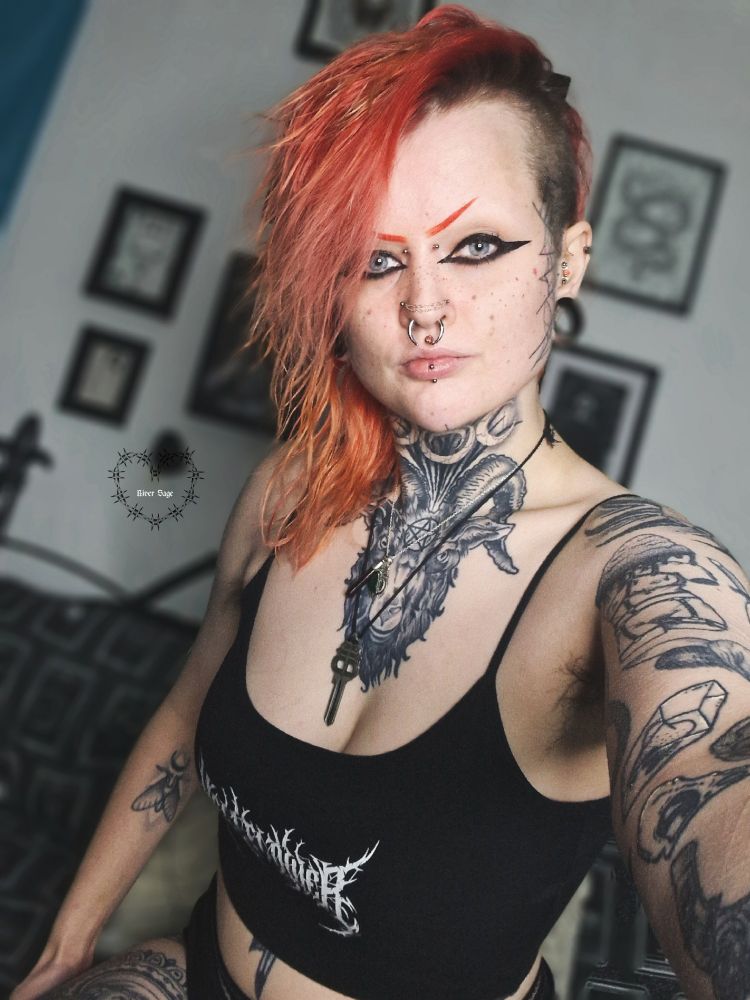 River is taking a selfie. They have their red hair over to one side, with two shaved sides. They are also wearing a Hellflower brand crop top, a key necklace and a blood vial necklace. They have eyeliner on and red eyebrows.