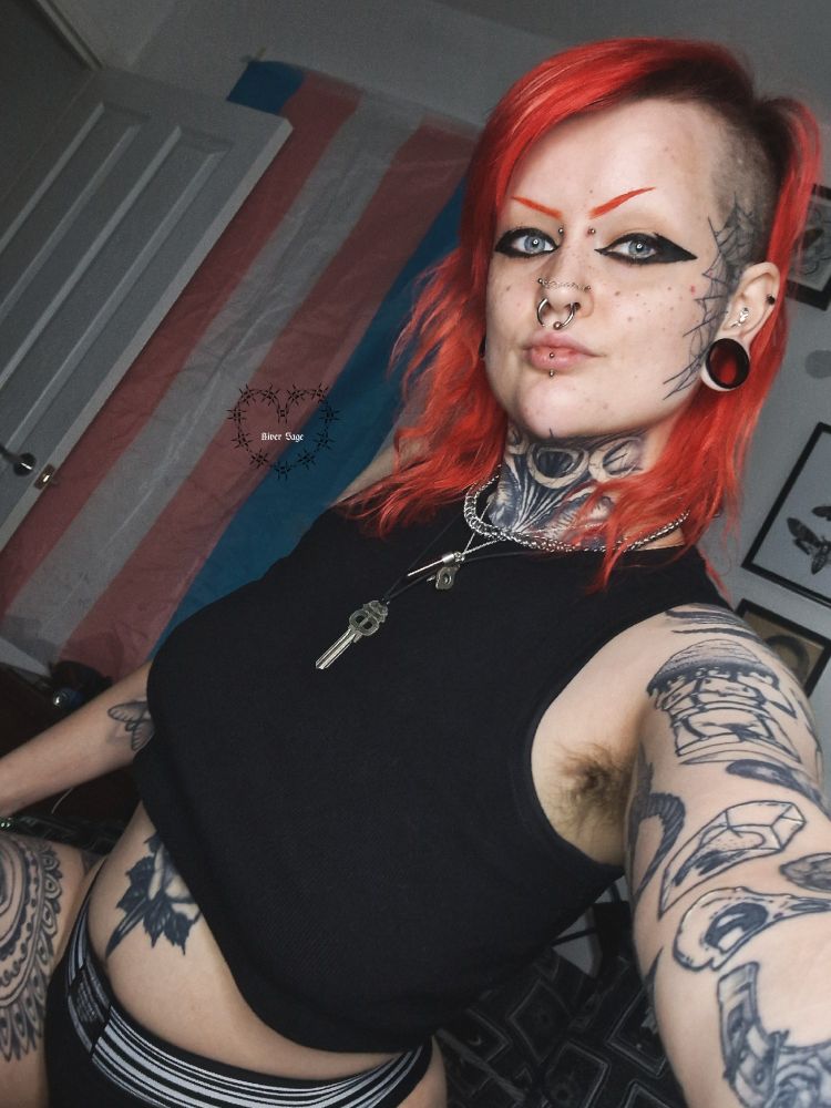 River is taking a selfie. They are wearing a black cropped tank top, with a black and white jockstrap. Their hair is red and to the side, and they are wearing black eyeliner. They have a key, a chain and a blood vial around their neck.