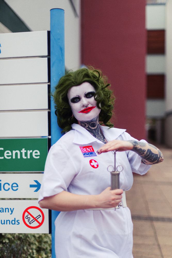 River is stood in front of a hospital sign, holding a metal syringe. They are dressed as Heath Ledger's Joker, in a hospital dress, a green wig, a white painted face, black eye makeup and red Chelsea grin style lipstick.
