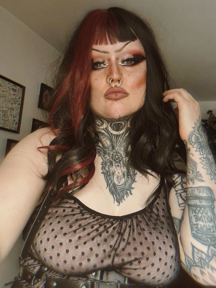 River is taking a selfie. They are in a drag look; they have a shoulder length black and purple wig on, and a dusty pink and black dress. They have bold makeup on, that is purple and pink. One hand is raised towards their face.