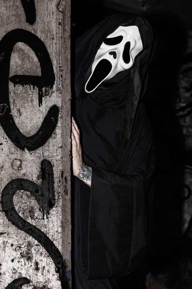 River is wearing a ghostface from Scream mask, with a long black robe on. They have their hand resting on the side of a pillar, with their head tilted to the side. They are in an abandon building with graffiti in the background.