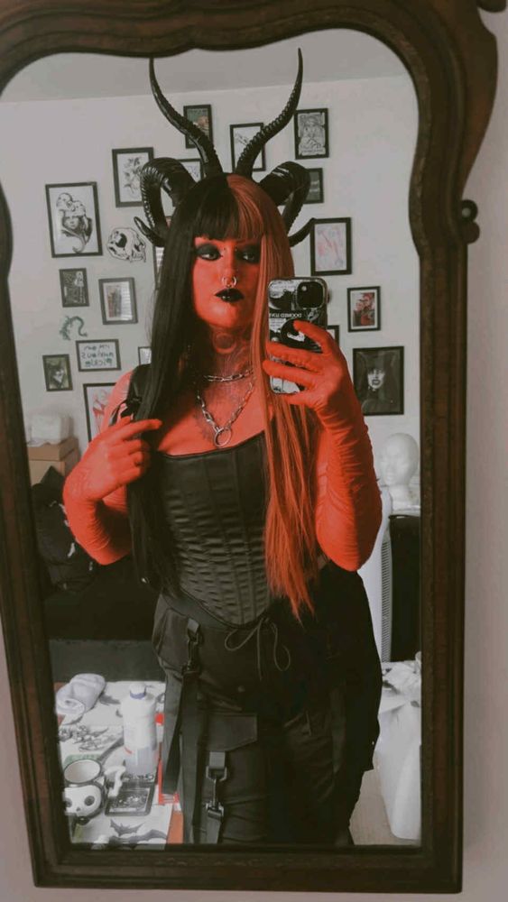 River is taking a mirror selfie, and the image is cropped to their thighs. They are wearing a Tiefling cosplay, where they are painted red and is wearing black horns. They have a black corset and harem style pants. 