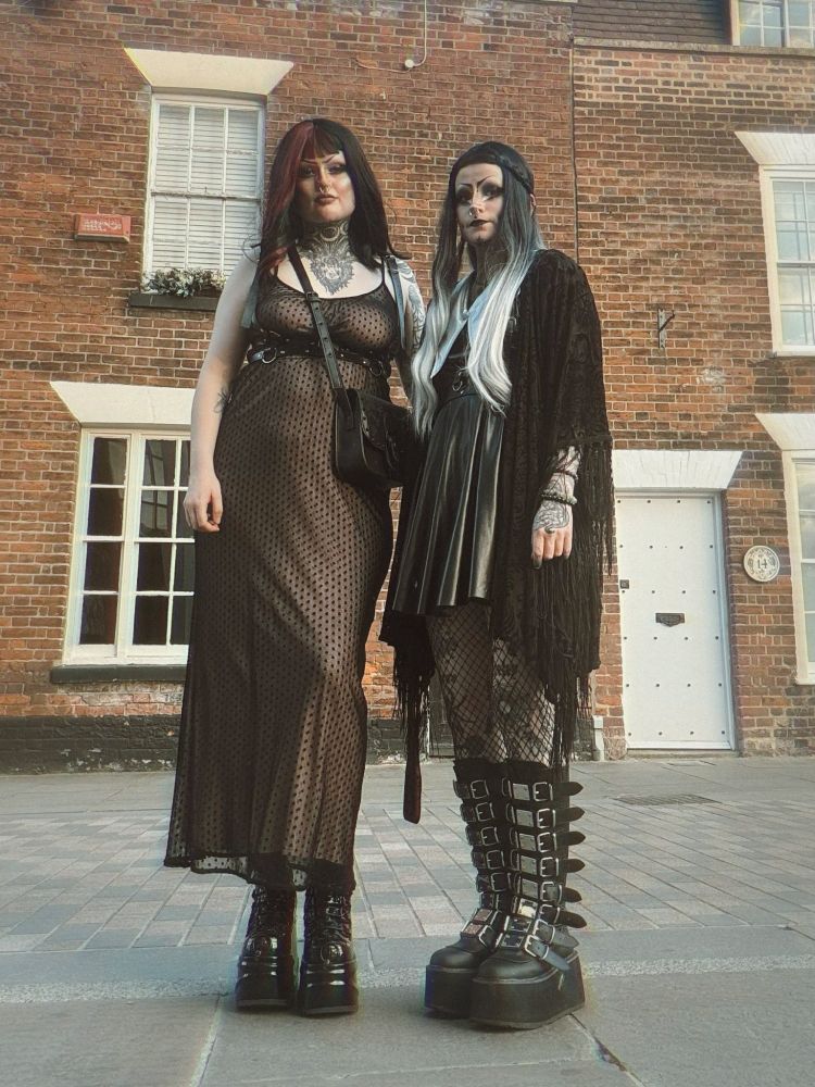 River and River are stood next to each other. River on the left is wearing a dusty pink and black dress, with a harness on and a shoulder bag. They also have big black platform boots. River on the right is wearing a black dress with a black kimono and big platform boots that goes to their knees. They have a black and silver wig on. Both of the River's have dramatic drag style makeup.