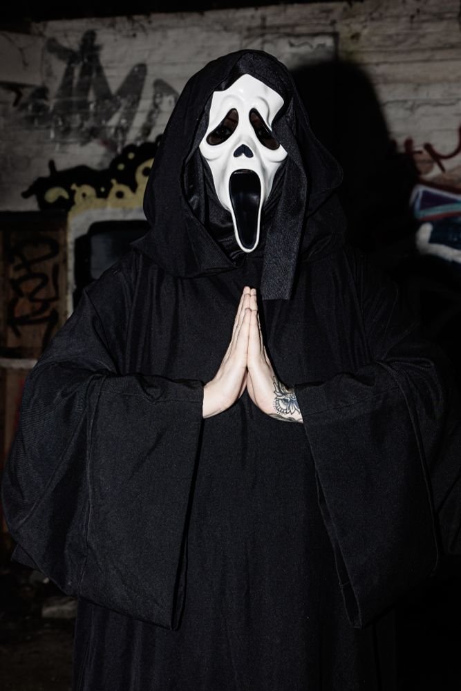 River is wearing a ghostface from Scream mask, with a long black robe on. They have their hands up to their chest with their hands in prayer. They are in an abandon building with graffiti in the background.