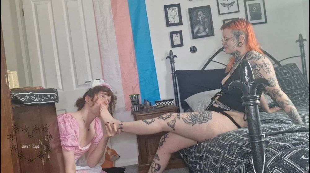 River is sitting on the edge of their bed, with their legs stretched out to their submissive. Their submissive is holding their foot up, with their toes in her mouth. River is getting their feet thoroughly worshipped. 
River is wearing a black corset style top and a back and white jockstrap. Their submissive, Juno, is wearing a cute pink maids outfit with a headband on.