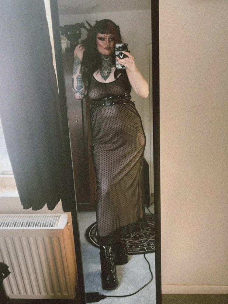 River is taking a mirror selfie. They have a short black and purple wig on, a dusty pink and black dress, a black harness and black platform boots. They have one hand raised towards their face.