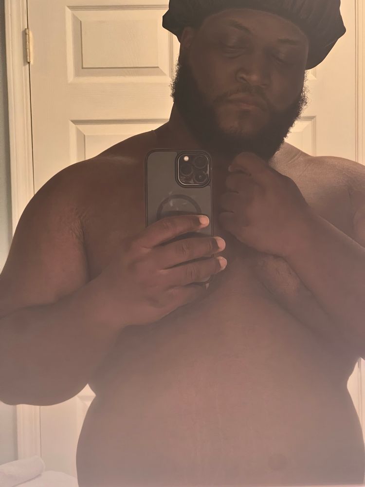 Fat black man showing off his upper torso in a black shower cap in front of a bathroom mirror