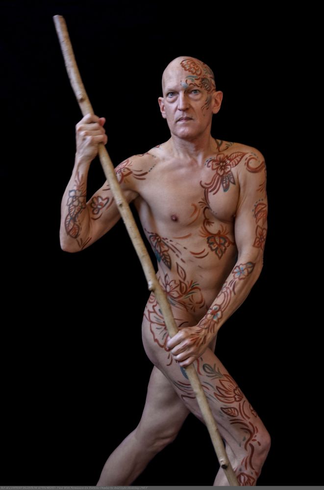 Color image of a nude male model in modest pose with a tall staff. Model is decorated with a bodypaint line design inspired by henna art, but made larger with body paint in order for the design to show up during online life modeling. 