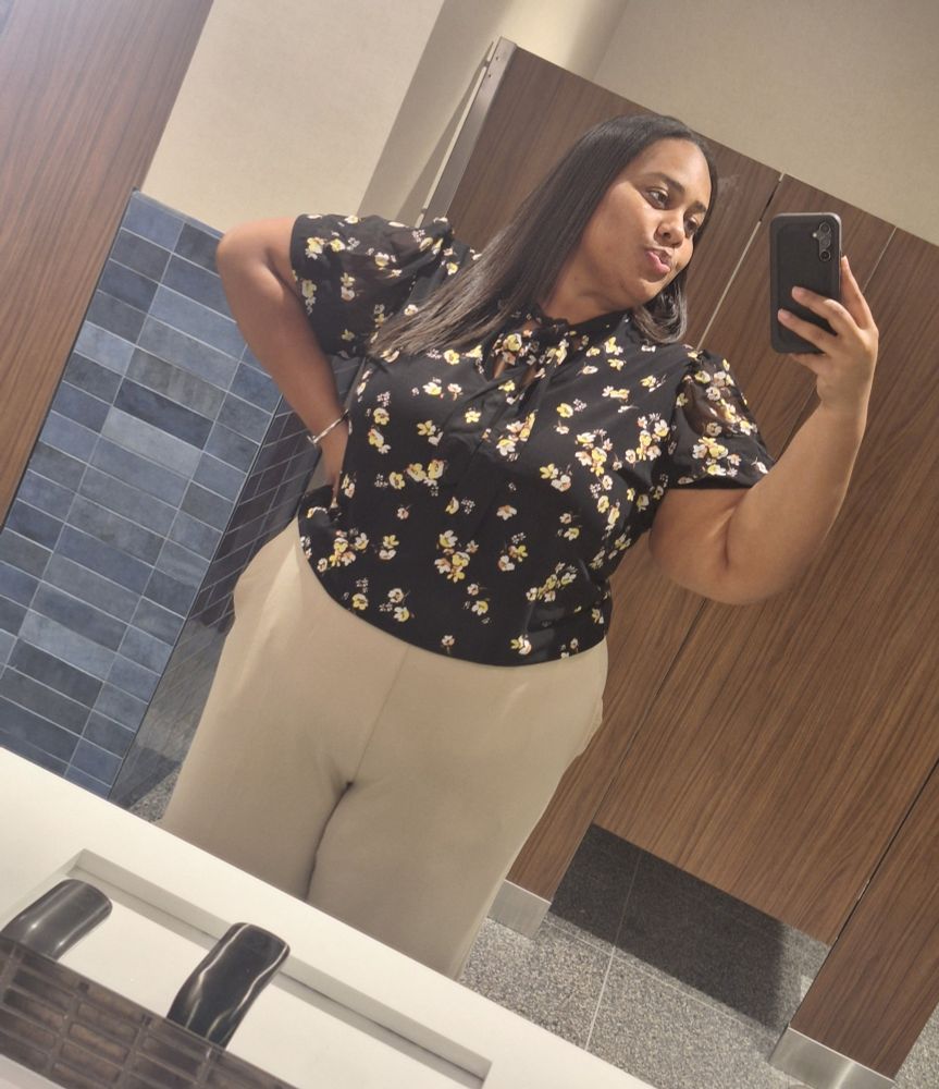 Black girl in a black shirt with small flowers on it and beige slacks takes a selfie from the knees up in a mirror. 