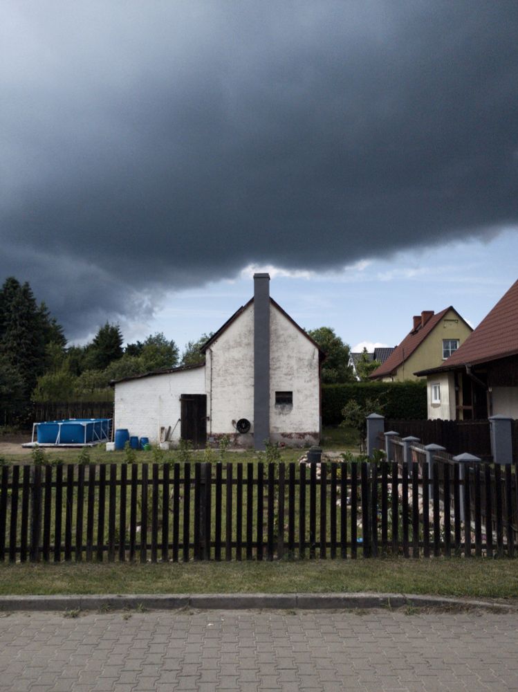 Biały dom za płotkiem z szarym kominem. Na niebie chmury które sprawiają wrażenie jakby wychodziły z komina. Po lewej basen a po prawej inne domy.