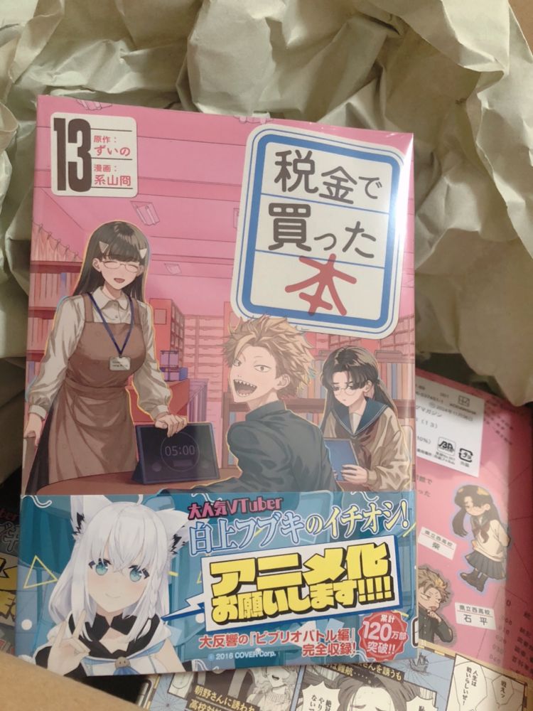漫画「税金で買った本」13巻単行本　原作者の家に届いたものを撮影した写真　帯はVtuberの白上フブキさんのイラストと「アニメ化お願いします！！！！」のコメントつき