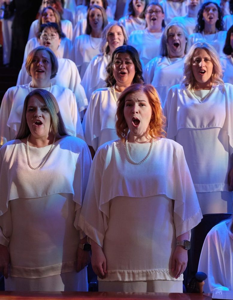 The women’s section of the Mormon Tabernacle Choir performs, wearing expressions of strained religious ecstasy. They’re wearing multi-layered white outfits that I will attempt to describe but will not be able to do justice. They sort of resemble stereotypical choir robes at the shoulders and chest, with boat necklines and wide sleeves, except they appear to be made of jersey and are abruptly cropped just below the bust with an extremely narrow, puckered hem. The effect is as if someone found a row of robes on hangers and hurriedly and maliciously took shears to the bottom two thirds, like a prank by mean high school girls. The underlayer is tunic-length and tight across the stomach and hips, pulled down over a straight long skirt in what seems to be the same material. The sleeves and underlayer hem are trimmed with the messiest, wrinkliest inch and a half of shiny white satin you’ve ever seen in your life. Sewing machines were definitely harmed in the making of this photo. The sleeves are bracelet length-ish, showing Apple watches and varying amounts of arm. Each ensemble is finished with a pearl necklace, which pools unevenly in the folds and creases in the outer robe. 