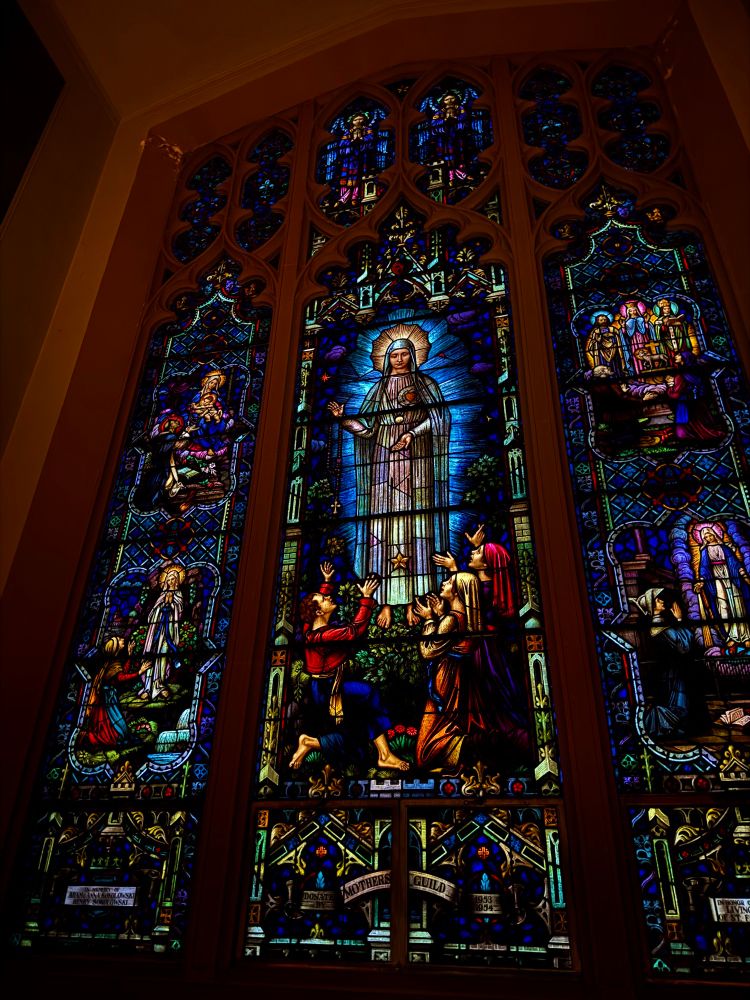 A tall, brilliantly jewel-toned stained glass window glows in early dawn light. A bright blue glow around Mary is especially vibrant. 