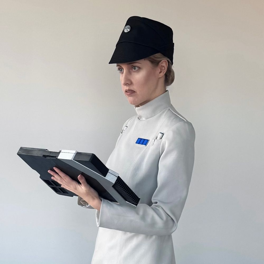A person dressed as Dedra Meero, from Andor, wearing their pale grey tunic and looking thoughtful and dour while holding a datapad. 