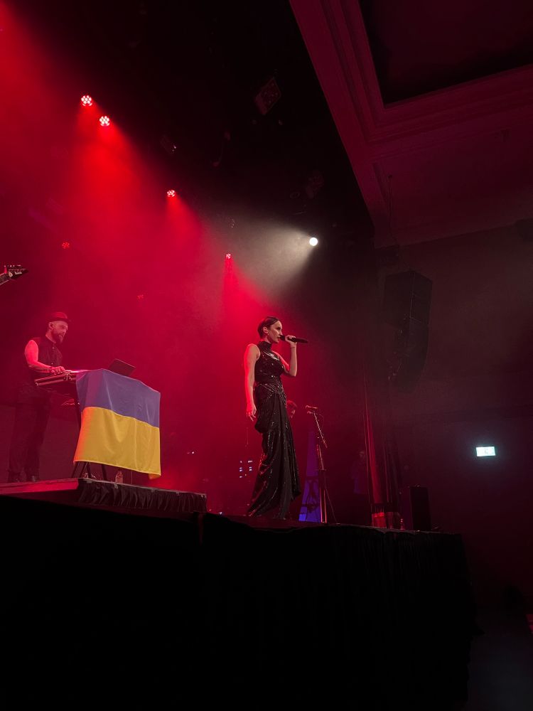 The band Go A performs on a small auditorium stage. Kateryna Pavlenko is in front wearing her signature Matrix-style slicked-back hair and layered leather outfit, singing intensely into her handheld mic. Taras Shevchenko wears a vest and a bowler hat and has his keyboard draped in a Ukrainian flag. The stage is foggy and dramatically lit in deep reds. 