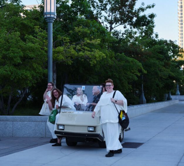 A group of people in their aforementioned dresses get cheerfully out of a golf cart on the Temple Square grounds. The full outfit is visible, with white skirts reaching almost to the ground and closed-toed black shoes sticking out. 