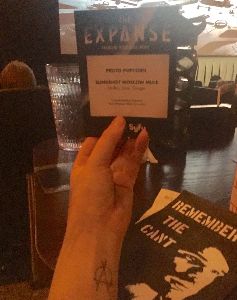A menu for the Expanse Season 2 premiere’s food specials is held up in front of a movie screen. There’s a scratchy OPA split circle tattoo on the arm holding it, and a Remember the Cant flyer featuring James Holden’s face on the table. 
