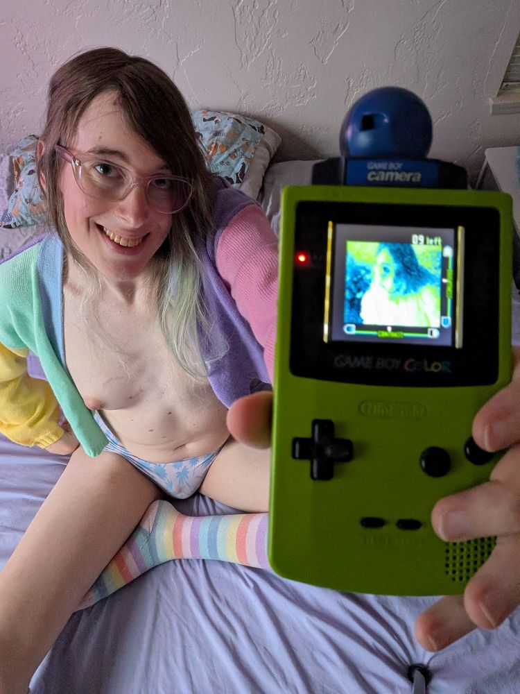 Photo of a smiling trans girl with her breasts exposed in a pastel sweater, holding up a Game Boy Color to take a selfie with a Game Boy Camera