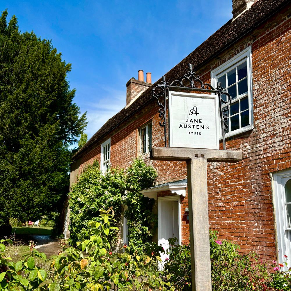 A red brick 18th century (?) English cottage with flowery vines climbing up the side. A sign in front of it reads “Jane Austen’s House”