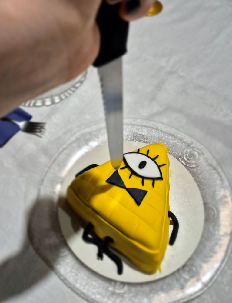 Mão segurando faca apontando pra um mini bolo no formato do Bill Cipher
