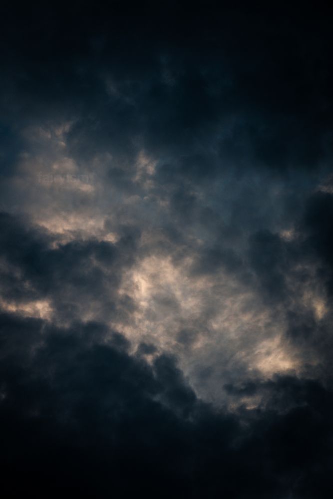 Very dark vertical photograph of the blue sky with dark and heavy clouds in it. Only in the center of the photo is some light, due to the sun behind the clouds. The top and the bottom of the frame are entirely dark. 