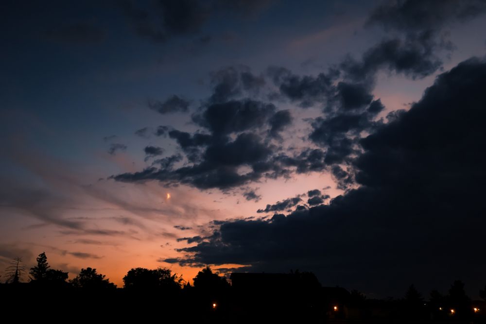 Photograph of the orange and dark blue sunset sky, with the silhouettes of trees and houses at the bottom of the photo. There is also the small crescent moon in the cloudy sky.