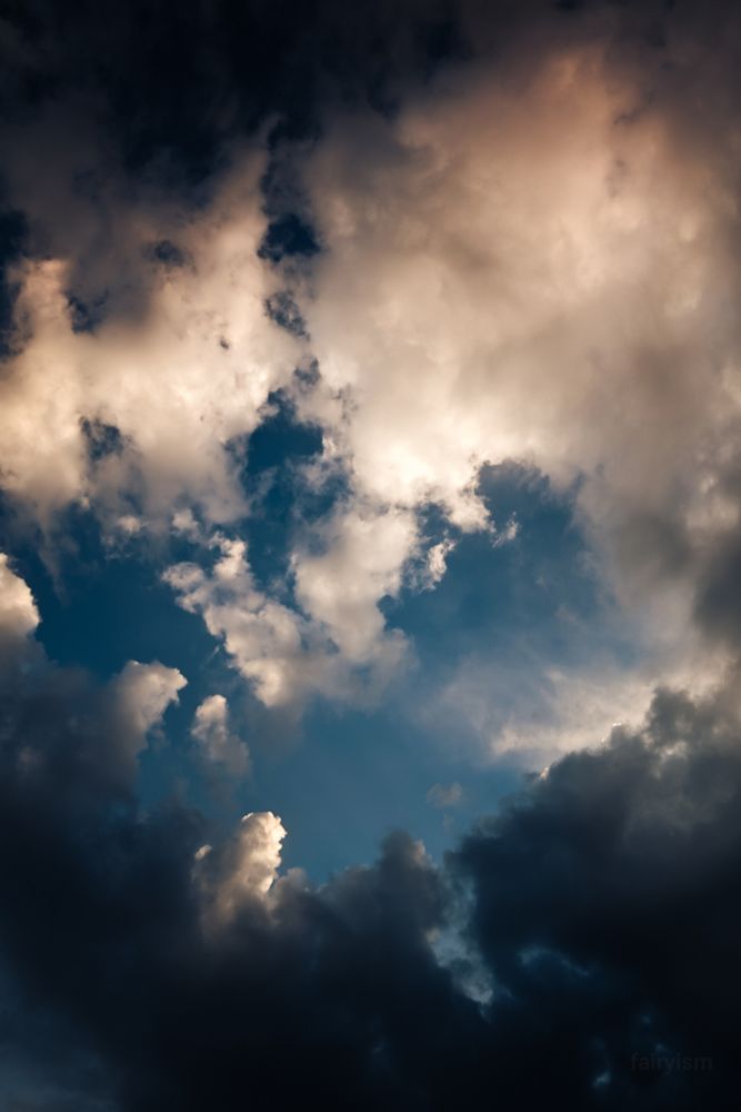 Vertical photograph of the blue sky filled with clouds during sunset time. In the bottom are darker clouds, while in the middle are some clouds which are more illuminated. In between is the blue sky peeking through. 