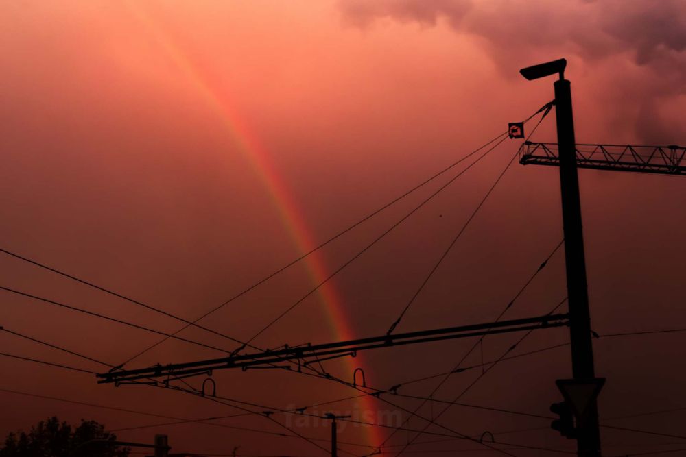 Photograph of several power lines from a tramway line. In the left of the photo is a power pole, connecting the chords. In the middle of the photo is a soft rainbow, radiating through the lines. The toning of the photo is quite orange / red / warm.