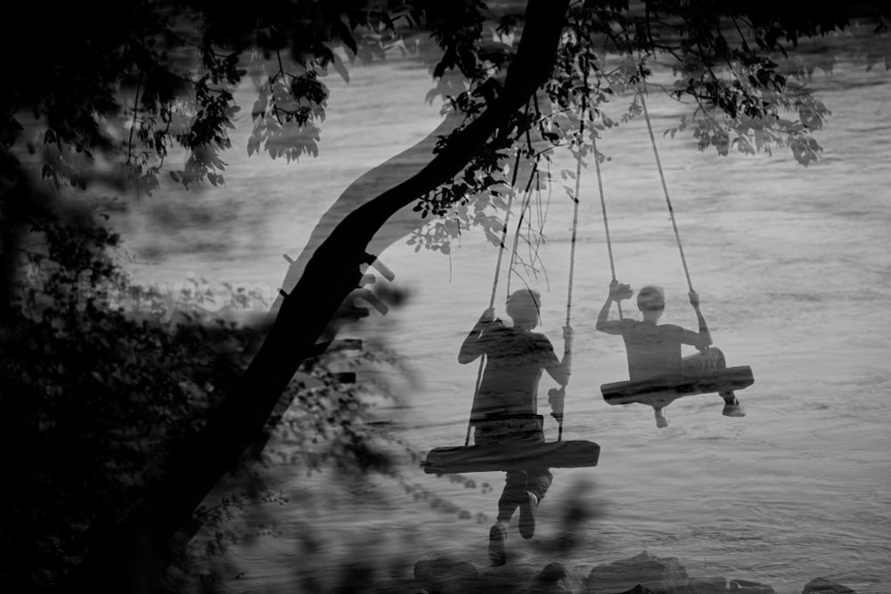 Black and white double exposed photograph of a person swinging on a swing, hanging from a thick branch of a tree, with the water of a river in the background. The person is two times in the frame, which is caused by the double exposure. From the left side are the silhouettes of leaves looming into the photo. 