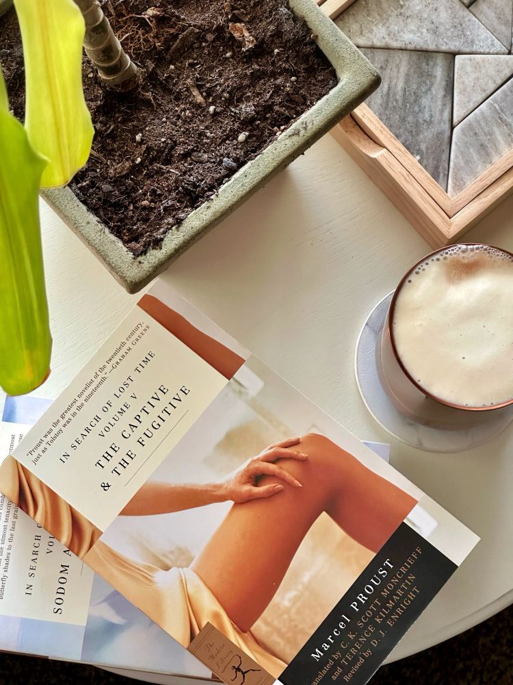 My side table, showing two books: The Modern Library edition of The Captive & The Fugitive sits on top of Sodom and Gomorrah. Also on the table, a hot drink with some froth, a table puzzle, and a house plant. 