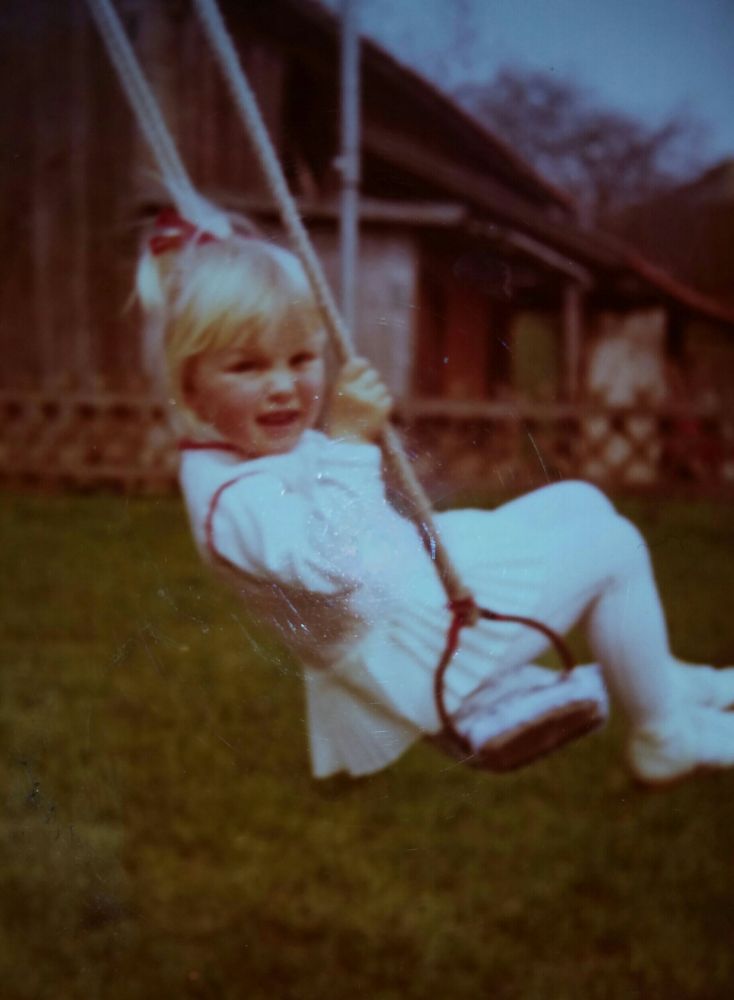 Die Userin sitzt in einem weißen Kleidchen und mit roter Haarschleife auf einer Holzschaukel und grinst in die Kamera.
