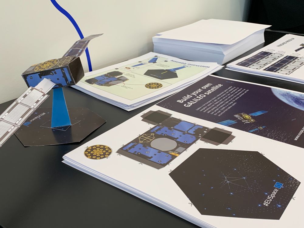 A paper model of the Galileo satellite displayed on a black table, next to printed cutout sheets and assembly instructions for building the satellite. The sheets include diagrams, folding guides, and the EU Space logo.