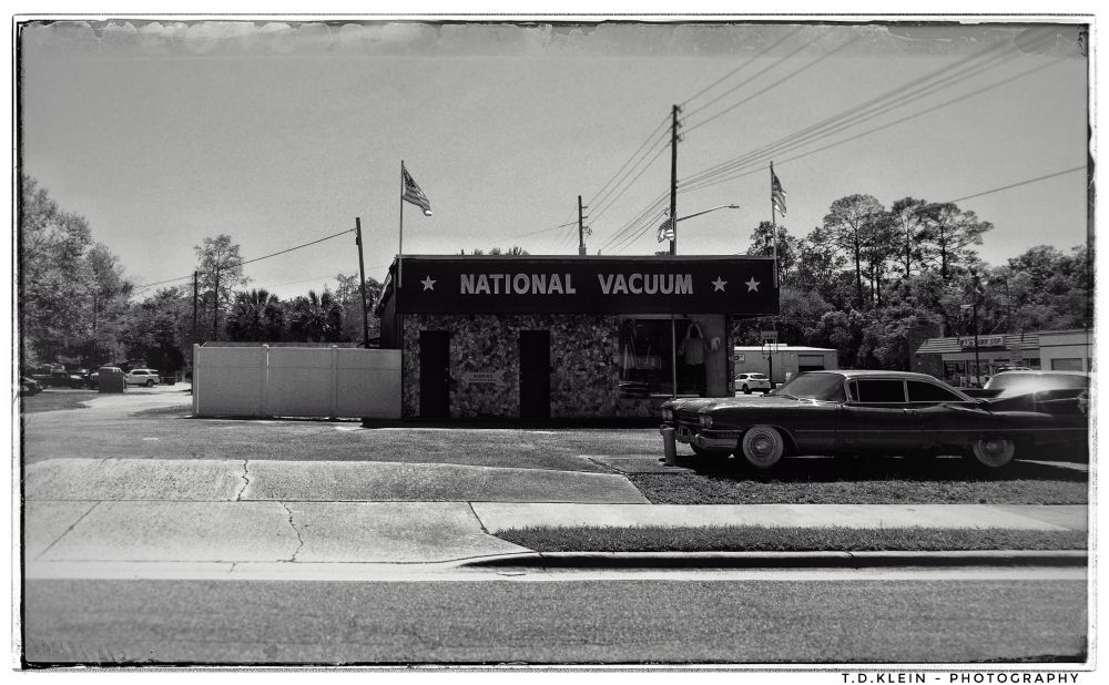 b/w picture of a vacuum cleaner shop near an eerily empty road, some old car parked in front of it - lots of patriotic Flags, 'National Vacuum' written above the front. Ironic.