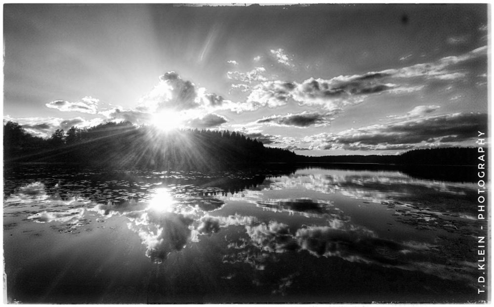 b/w photography of a summer sunset mirrored by a silent lake, forest and some clouds create s solemn atmosphere 