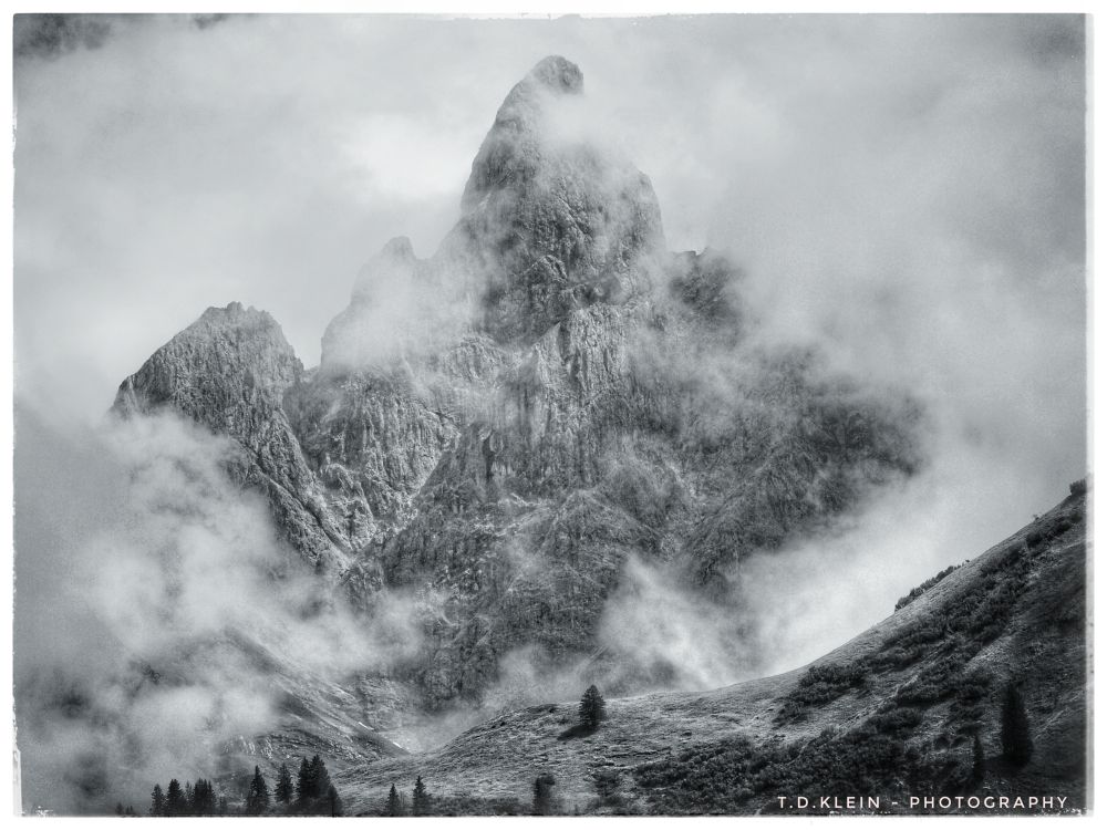b/w pic of an imposing, spectacular mountain, briefly showing its rugged, jagged face between a layer of clouds, steep, crenelated Dolomite rock ignited by the sun - in the foreground steep alpine meadows, some trees, Oberallgäu, Germany, 2021