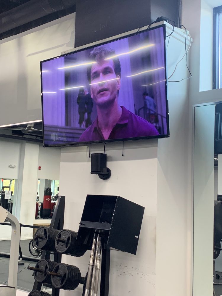 a TV at a gym above a rack of weights showing Patrick Swayze in the movie “Ghost”