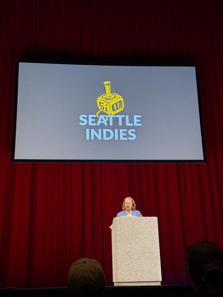 A speaker on stage speaking. He is in front of a red curtain