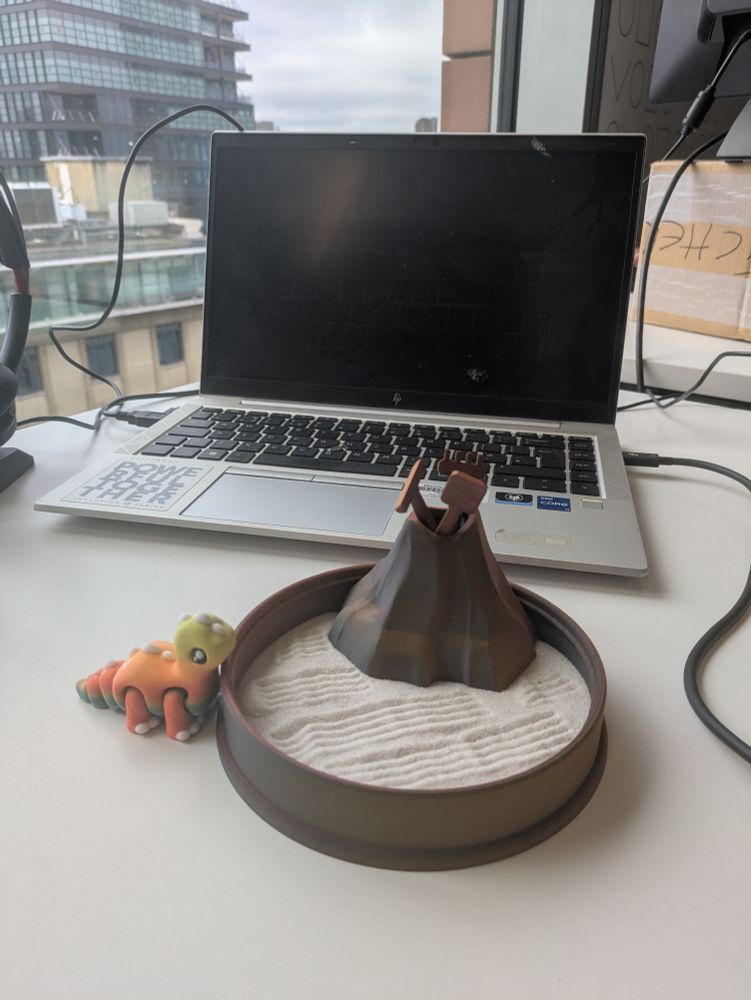 A desk toy sand garden sitting on a white desk, with a Dino approaching on the desk. Behind, part of the desk toy, is a mountain which contains sand tools. Behind that, out of focus, are normal desk things: a laptop and a headset. The Dino is coloured yellow at the top through orange and the green on its belly.