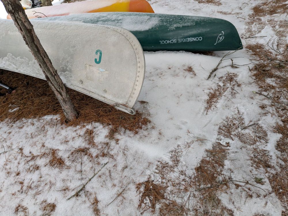 A trio of canoes covered in snow. The furthest back is yellow-orange with no visible writing, the one i the middle ground is darker green with the words "Conserve School" written in white, and the one in the foreground is white with a green "C" on it.