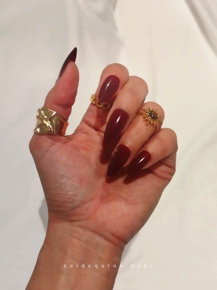 Asian FinGod Zeldaquine's left hand against the white sheets palm-facing showing her burgundy stiletto nails, her fingers ornamented with gold rings