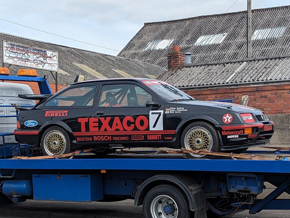 Photo shows a Ford (Sierra) Cosworth RS500, in black and red Texaco racing livery on the  back of a flatbed truck. 